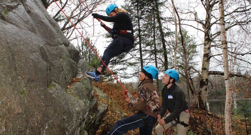 Three people wearing safety gear participate in rock climbing during the family seminar of an outward bound intercept course.